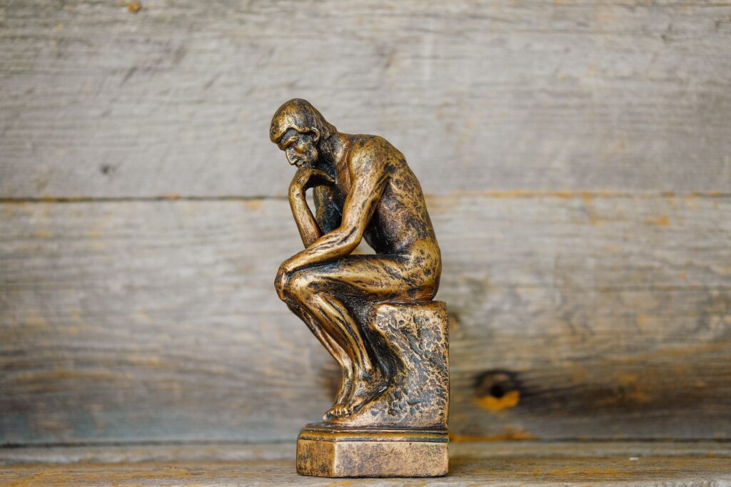 A small, bronze statue of a man sitting the the "thinker" pose. He is sitting, hunched over, his elbow resting in his lap, his chin resting on his hand.