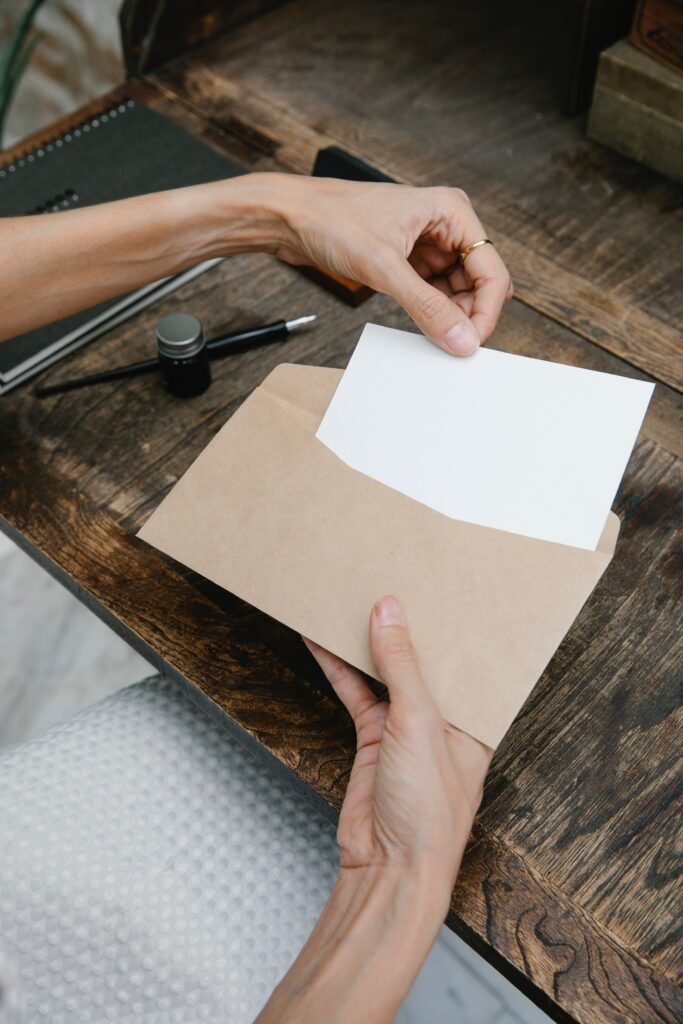 An image of a blank piece of paper being placed in a brown paper.. There is a desk in the background with writing utensils in the background.