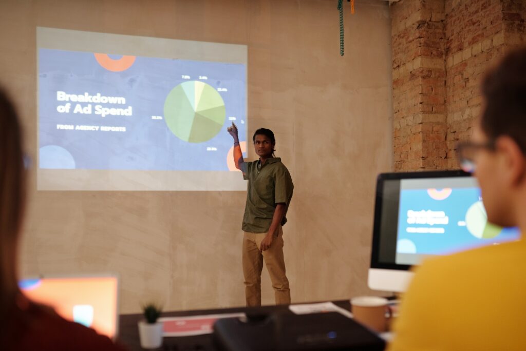 A photograph of a man standing in front of a projected power-point presentation. He appears to be presenting to a small group of work colleagues. 