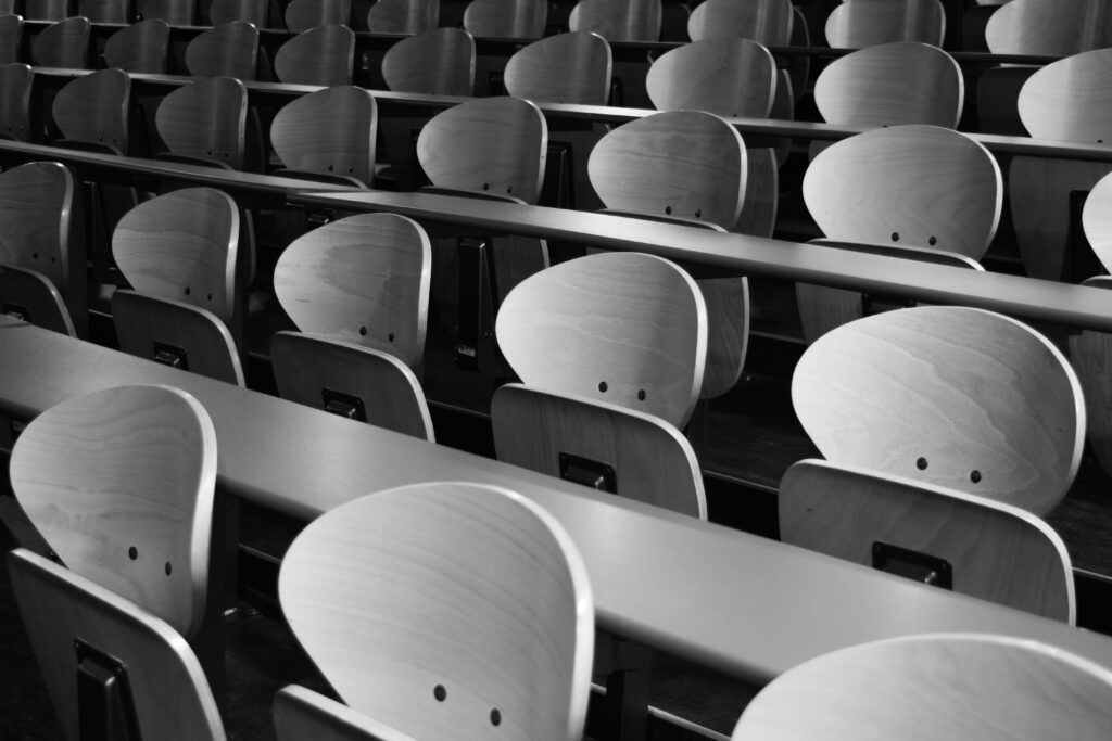 A black and white photograph of rows of wooden seats behind long desks. The desk formation resembles  that of a university lecture hall.
