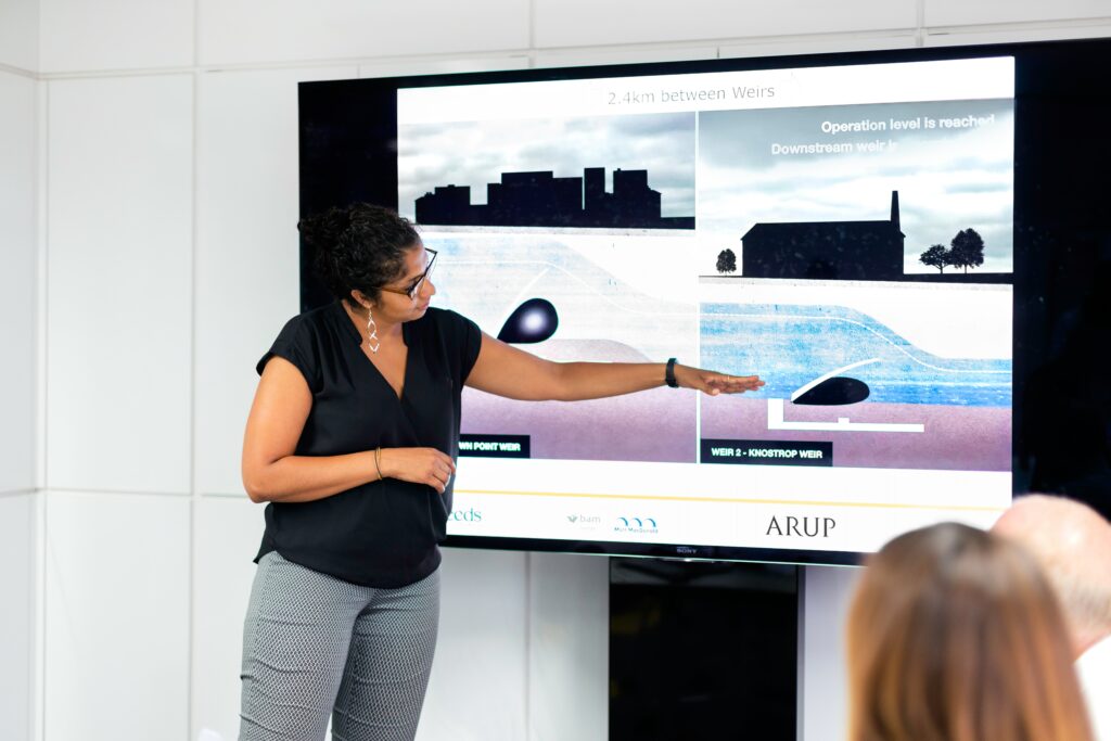 An image of a woman giving a presentation. She is standing in front of what appears to be a smart board, and motioning to one of four images on the screen.
