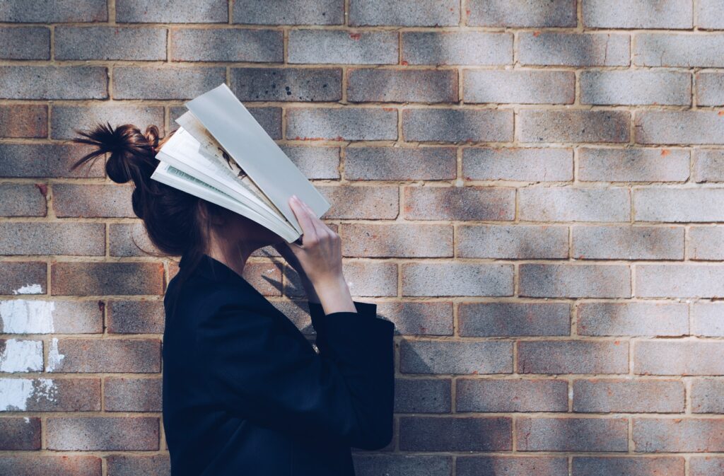 A photograph of a woman standing in front of a brick wall. The woman is photographed from the chest, up, and only her profile is visible. She is holding a book open, and has plonked it down on her face, hiding her face from view.