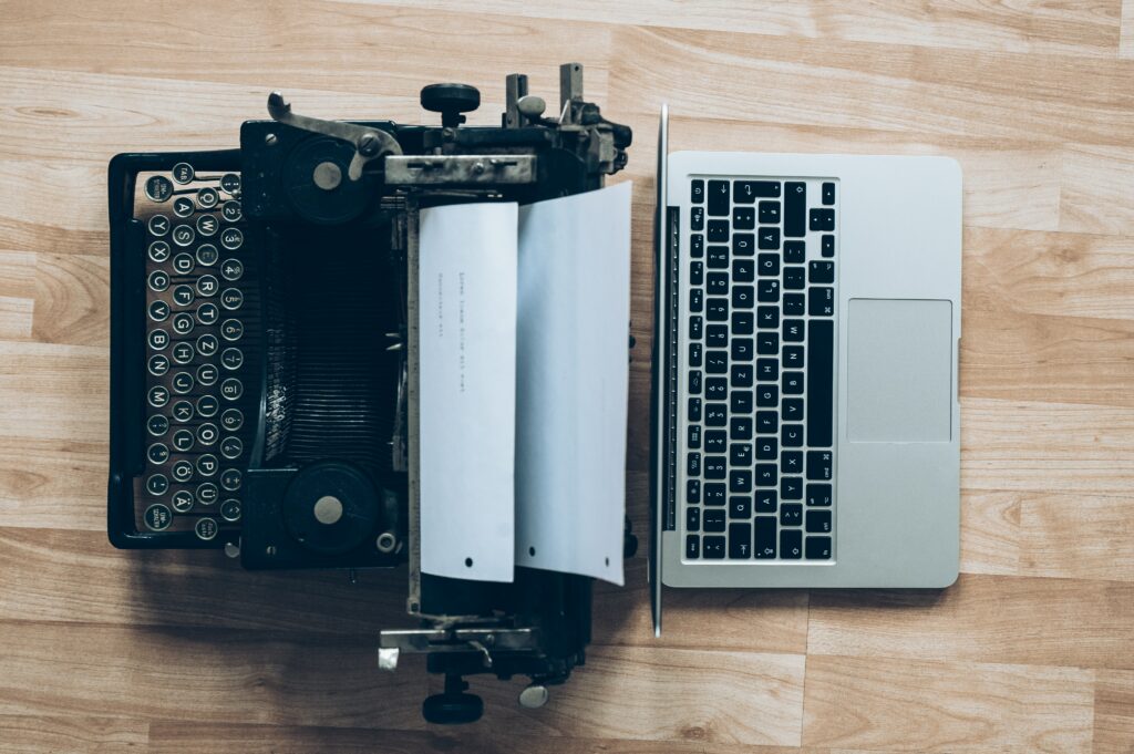 Top-down photograph of a typewriter and an open laptop. The two devices are back-to-back with one another.