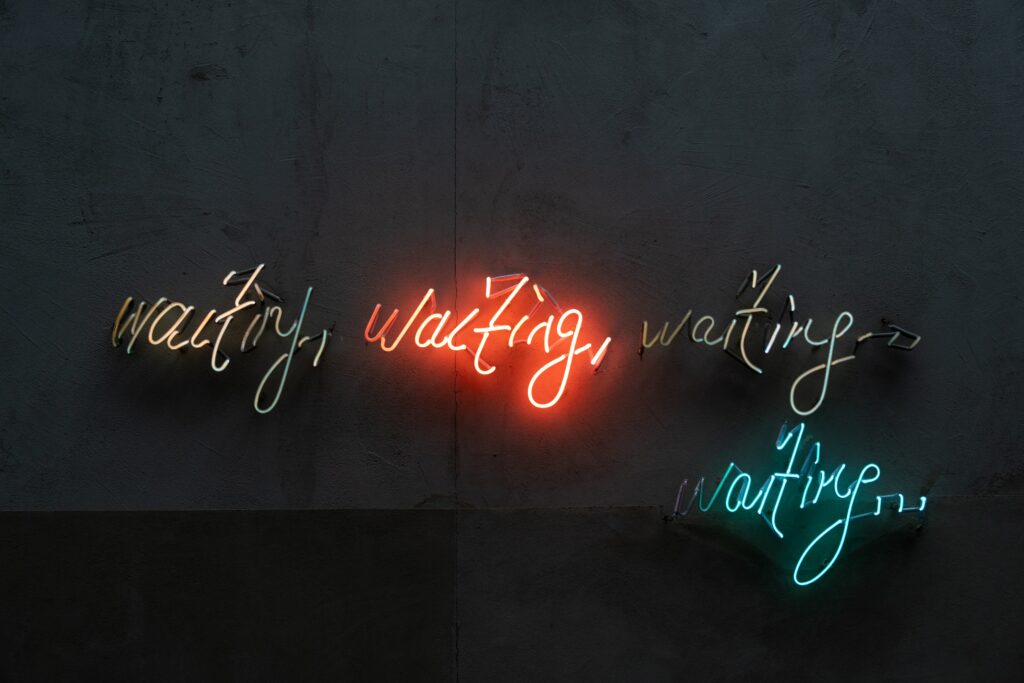 A photograph of neon-lights in the dark, which write out, "waiting, waiting, waiting... waiting..."