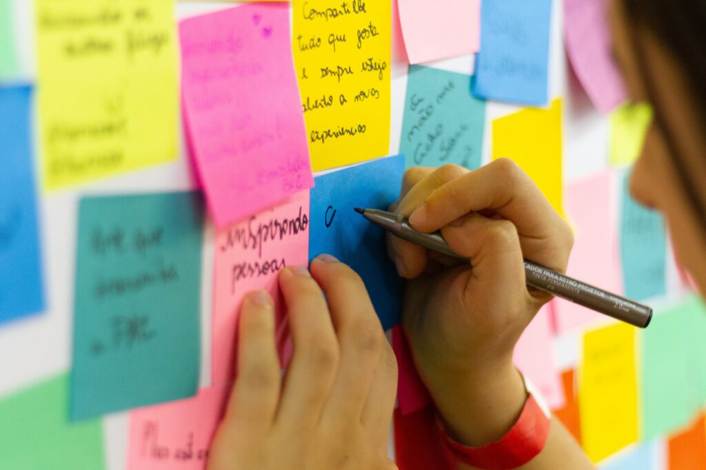 A close-up photograph of wall covered in filled-out sticky notes. A woman is posed as she begins to write on a blank note.