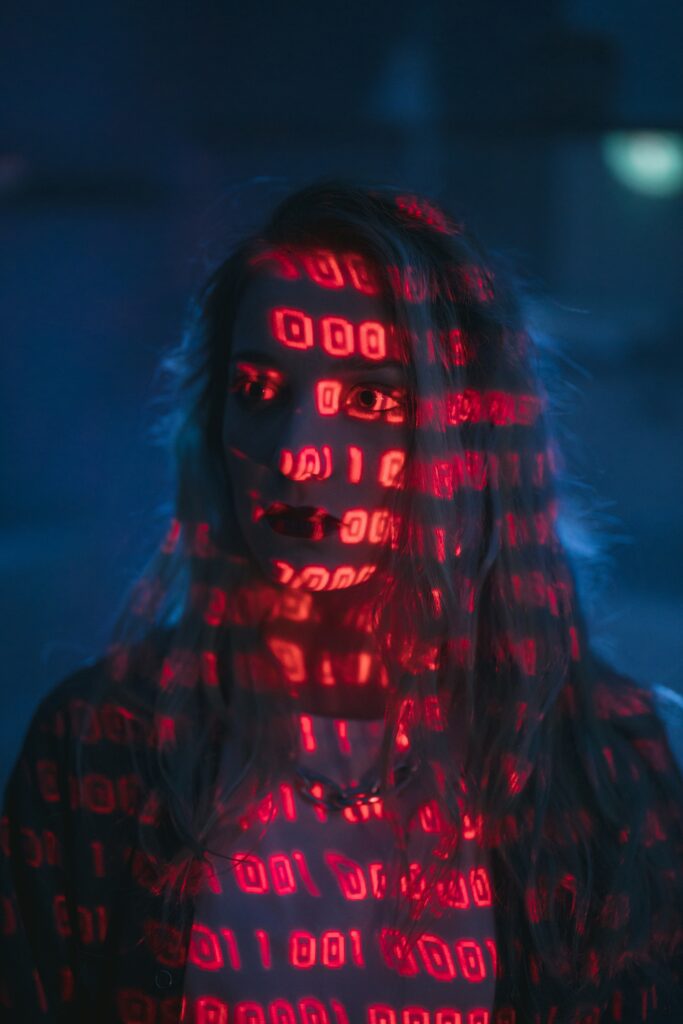 A moody, darkly lit photograph of a woman. She is looking to the side, and has a striking projection of red binary code projected across her face.